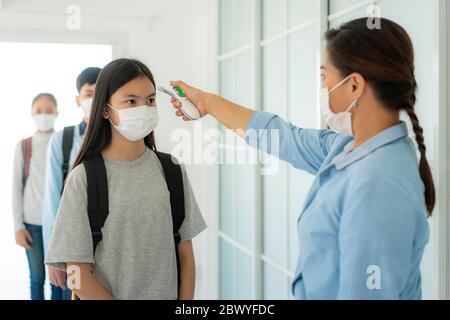 Insegnante di donna asiatica che usa termometro temperatura di screening studente per la febbre contro la diffusione di COVID-19, mentre studente tornare a scuola, nuovo Foto Stock