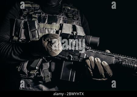 Foto di un soldato in guanti tattici e gilet con armatura in piedi e ricaricando fucile da cecchino su sfondo nero. Foto Stock