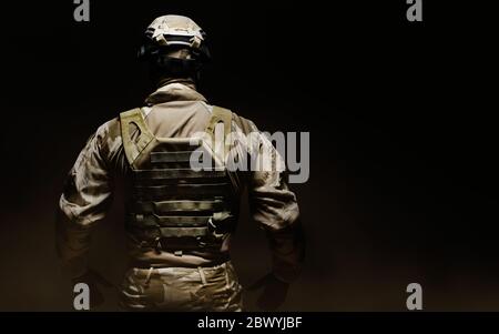 Foto di un soldato camuffato nel deserto completamente attrezzato in maschera, casco, armatura e guanti su sfondo nero nebbia vista posteriore. Foto Stock