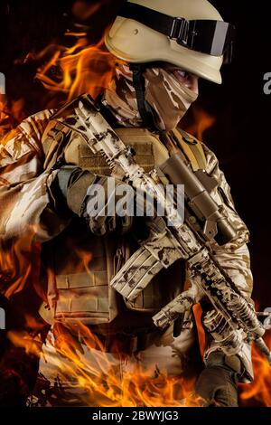 Foto di un soldato completamente attrezzato in uniforme camuffata, casco, guanti e armatura che tiene il fucile su sfondo fuoco sparente. Foto Stock