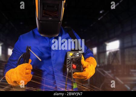 Foto di saldatore in uniforme, casco e guanti che tengono inverter ed elettrodi su sfondo industriale. Foto Stock