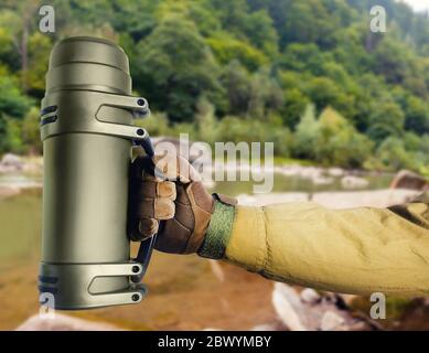 Foto di soldato mano in guanto che tiene thermos tattici su sfondo natura. Foto Stock