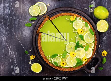 Cheesecake con fette di lime e limone, menta fresca, frutta candita e fiori di pansy su fondo di pietra scuro. Vista dall'alto. Foto Stock