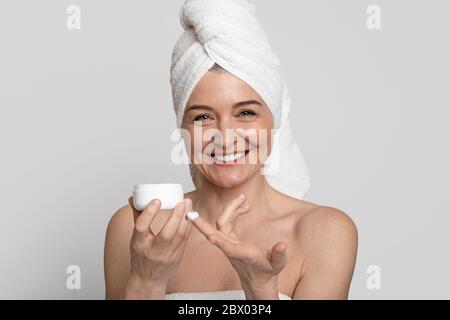 Donna matura positiva con asciugamano sul vaso di sostegno della testa con crema idratante Foto Stock