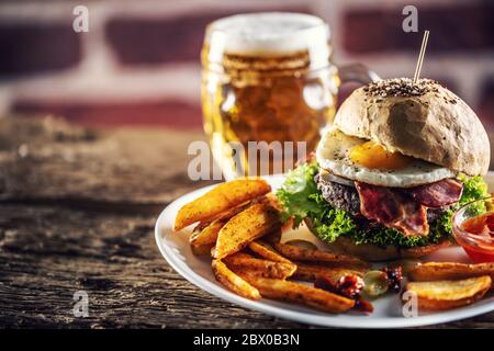 Hamburger con un panino di sesamo, uova fritte e pancetta, insalata e spicchi di patate con una birra alla spina sullo sfondo Foto Stock