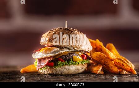 Hamburger di manzo con uova fritte, pancetta, formaggio, lattuga e peperoni in un panino di sesamo con spicchi di patate e paprika affumicata Foto Stock