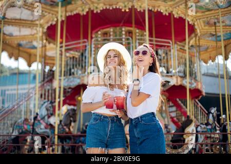 due giovani ragazze alla moda parlano e ridono. tenete i cocktail in mano. Vacanze nel parco impazzire insieme, con occhiali da sole a specchio, vista incredibile o Foto Stock