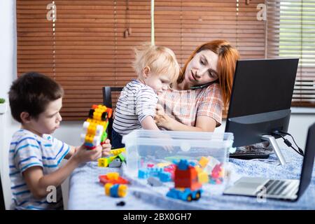 Una giovane mamma donna che parla al telefono e che cerca di lavorare in un computer in un lavoro remoto durante il periodo di autoisolamento in connessione con la co Foto Stock