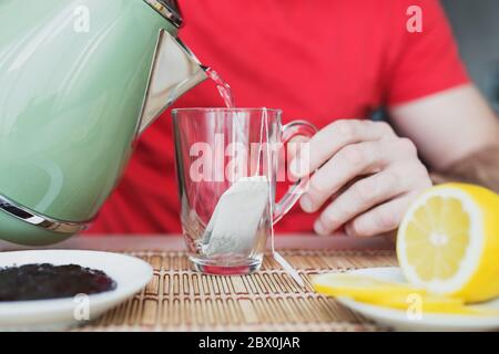 Uomo versando acqua bollente dal bollitore alla tazza di vetro per preparare il tè - colazione Foto Stock