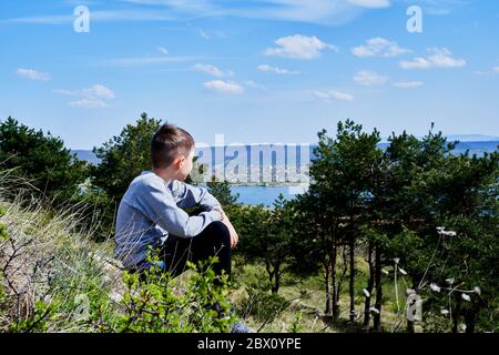 un ragazzo si siede su una collina e guarda lago Foto Stock