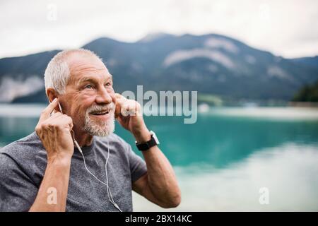 Uomo anziano con auricolari in piedi vicino al lago in natura, ascoltando musica. Foto Stock