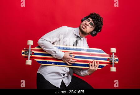 Giovane uomo con cuffie e longboard in uno studio su sfondo rosso. Foto Stock