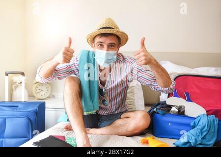 Felice uomo pronto a viaggiare in vacanza durante la stagione del coronavirus seduto su un letto con i pollici su ok e la sua valigia imballato Foto Stock