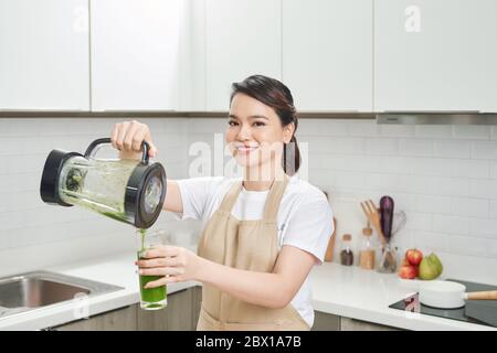 Giovane femmina in grembiule di lino che versa una bevanda verde frullato dal frullatore alla bottiglia circondata da verdure e verdure. Foto Stock