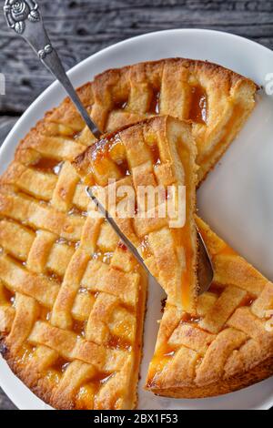 primo piano di torta di albicocche a fette con crosta di torta a graticcio che si stocca su una pala per torte vintage su un piatto bianco su un rustico tavolo di legno, vertica Foto Stock