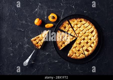 primo piano di torta di albicocche a fette con una crosta di torta a graticcio su un piatto nero, una fetta di torta su una pala di torta vintage su una linguetta di cemento Foto Stock