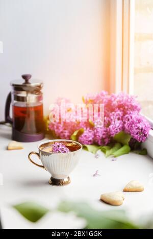 Tè caldo fresco in una bella tazza di porcellana con un bouquet lilla sullo sfondo su un davanzale bianco Foto Stock