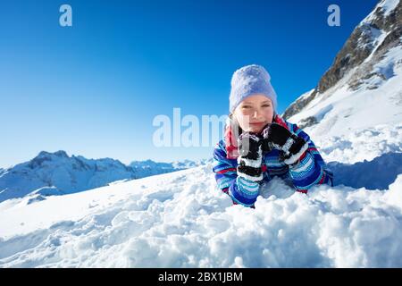 Felice ragazza sorridente si stese in neve che posa la testa sulle mani e sorridi indossando cappello invernale con la montagna sullo sfondo Foto Stock