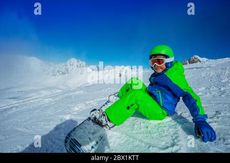 Happy boy su snowboard siediti sulla neve in casco e maschera con attrezzatura sportiva completa Foto Stock