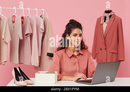 Giovane donna seduta al tavolo e con il laptop che acquista i vestiti online Foto Stock
