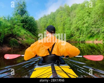 Gli uomini in un cappello e impermeabile di colore arancio galleggia su un kayak sulla foresta fiume tranquillo, il bellissimo paesaggio, un tempo mutevole, attivamente Foto Stock