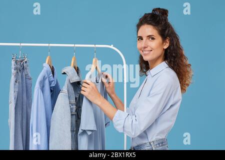 Ritratto di giovane donna sorridente alla macchina fotografica mentre sceglie la giacca di jeans sul rack nel negozio Foto Stock