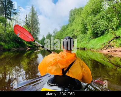 Gli uomini in un cappello e impermeabile di colore arancio galleggia su un kayak sulla foresta fiume tranquillo, il bellissimo paesaggio, un tempo mutevole, attivamente Foto Stock