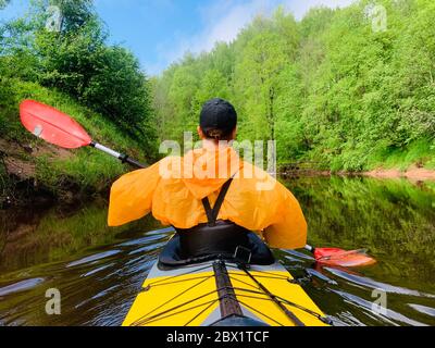 Gli uomini in un cappello e impermeabile di colore arancio galleggia su un kayak sulla foresta fiume tranquillo, il bellissimo paesaggio, un tempo mutevole, attivamente Foto Stock