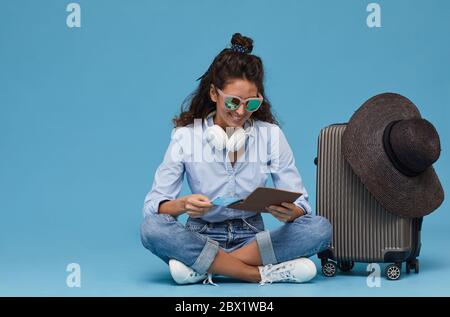 Giovane donna in occhiali da sole seduto sul pavimento vicino ai bagagli e utilizzando tablet digitale lei ordinare i biglietti on-line Foto Stock