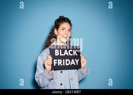 Ritratto di giovane bella donna che tiene il cartello del Black Friday e sorridendo alla macchina fotografica contro lo sfondo blu Foto Stock