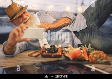 Uomo anziano sorridente mentre prende selfie con la fotocamera dello smartphone mobile - pensionato felice di divertirsi in barca festa bevendo champagne mangiare frutta Foto Stock