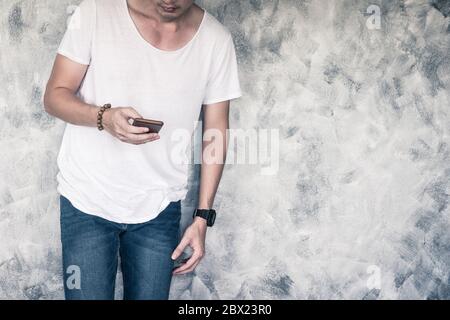 Giovane uomo che indossa una t-shirt bianca e jeans blu, che tiene uno smartphone e si trova su uno sfondo grigio. Spazio libero per il testo Foto Stock