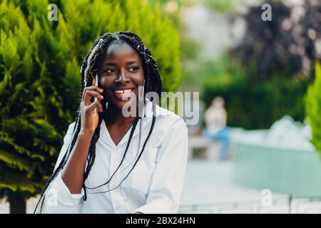 Primo piano ritratto di una giovane afroamericana sorridente con i pigtail seduti su una panchina all'aperto in una giornata di sole. Foto all'aperto. Foto Stock