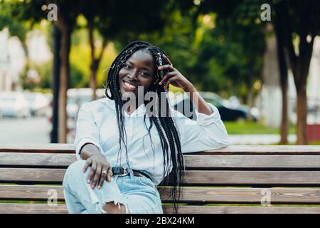 Primo piano ritratto di una giovane afroamericana sorridente con i pigtail seduti su una panchina all'aperto in una giornata di sole. Foto all'aperto. Foto Stock