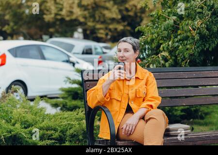 Donna anziana di tendenza, felice ed elegante, vestita nelle strade della città con un bicchiere di caffè in mano. Concetto anti-invecchiamento Foto Stock