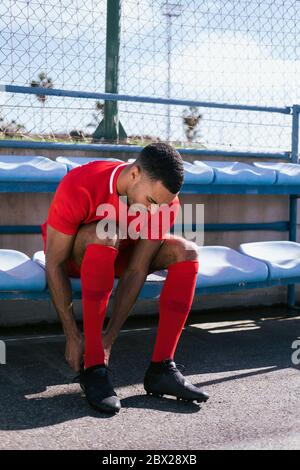 Giocatore di calcio africano americano maschio seduto e che fissa gli stivali banco Foto Stock