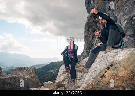 Concetto: Avventura. Arrampicatori uomo e donna con casco e imbracatura. Riposarsi su una roccia. Parlare e gesturarsi guardando l'orizzonte. Doi Foto Stock