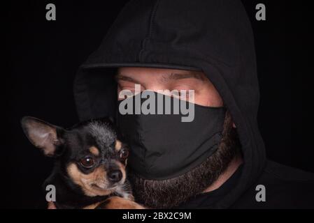 Uomo in una maschera nera, ragazzo mascherato in auto-isolamento con un cucciolo, ragazzo con cucciolo. Uomo in una maschera su sfondo nero con un cane. Chihuahua tra le braccia Foto Stock