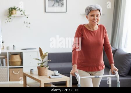 Ritratto di donna anziana che sorride alla fotocamera mentre usa il camminatore nella stanza a casa Foto Stock