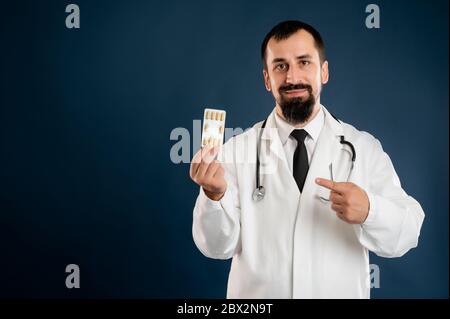 Ritratto di medico maschile con stetoscopio in uniforme medica mostrando pillole arancioni che si pongono su uno sfondo blu isolato. Foto Stock