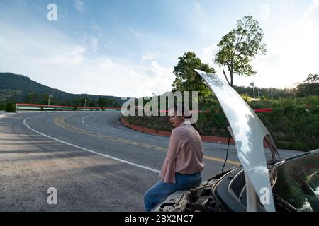 Una giovane bella signora seduta vicino alla macchina per chiedere aiuto sulla strada pubblica nella zona forestale a monte e cielo di fondo Foto Stock