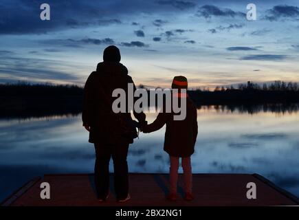 Silhouette di un adulto e di un bambino che si trova su un molo e guarda nel cielo serale Foto Stock