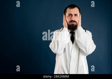 Ritratto di medico maschile con stetoscopio in uniforme medica che copre orecchie come il concetto sordo che si pone su uno sfondo blu isolato. Foto Stock