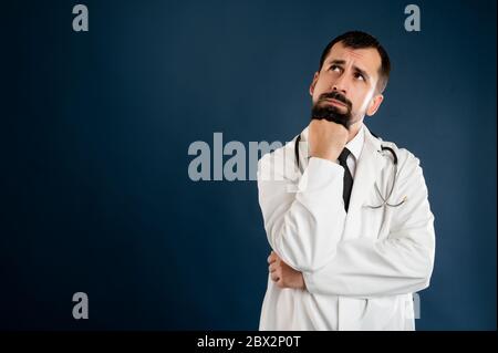 Ritratto di medico maschile con stetoscopio in uniforme medica tenendo la mano sotto il mento, avendo dubito di posa su uno sfondo blu isolato. Foto Stock