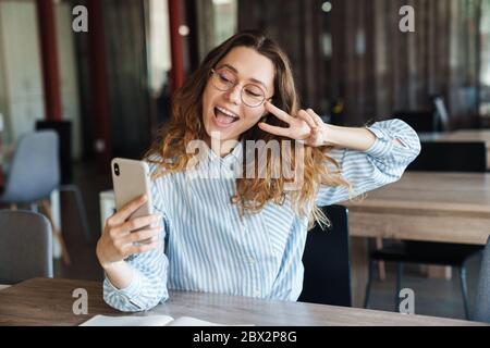 Immagine di una donna gioiosa bella gesturing segno di pace e scattare foto selfie sul cellulare mentre studia in classe Foto Stock