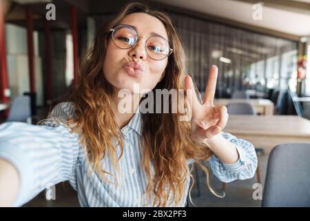 Immagine di felice bella donna gesturing segno di pace e scattando foto selfie in sala conferenze Foto Stock