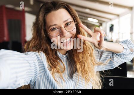 Immagine di felice bella donna gesturing segno di pace e scattando foto selfie in sala conferenze Foto Stock