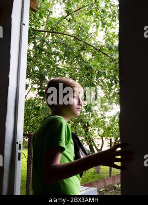 Ragazzo caucasico dai capelli biondi si trova nella porta di una casa contro un giardino Foto Stock