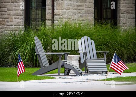 Due sedie grigie Adirondack seduta su un piccolo patio con tavolo tra loro di fronte a erba decorativa alta e finestre di casa di roccia - Fla americana Foto Stock