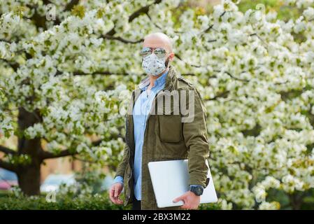 Un uomo calvo con una barba in una maschera medica per evitare il coronavirus diffusione passeggiate con un computer portatile nel parco. Un ragazzo indossa una maschera N95 e un pilota Foto Stock